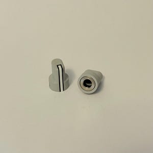 Op de afbeelding: Twee grijze bedieningsknoppen. Eén knop staat rechtop en toont een verticale zwarte streep met een witte rand. De andere knop ligt op zijn kant en onthult een zilverkleurig interieur. Beide knoppen liggen op een wit oppervlak.