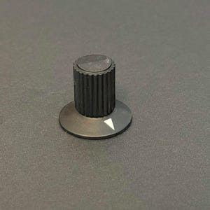 May include: A black control knob with a ridged cylindrical body and a circular base. The base features a white triangle indicating the knob's position. The knob is designed for adjusting settings on electronic devices.