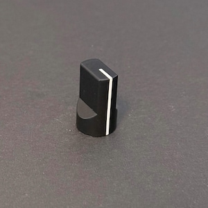 May include: A black control knob with a white stripe. The knob has a rectangular top and a curved base. The white stripe runs down the front of the knob. The knob is likely used for adjusting settings on electronic equipment.