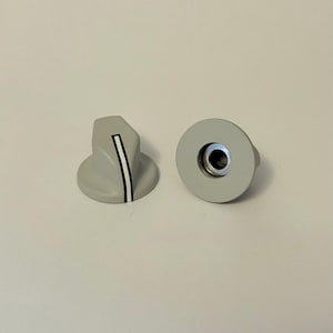 Puede incluir: Dos perillas de color gris claro. Una perilla es redondeada con una base circular y una inserción de metal. La otra perilla tiene forma de triángulo redondeado con una franja blanca. Estas perillas son probablemente para electrónica o electrodomésticos.