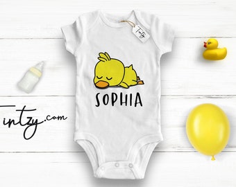 May include: White baby onesie with a yellow cartoon duck sleeping on it. The onesie has the name "SOPHIA" printed below the duck.