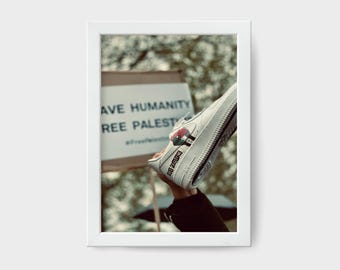 May include: A white sneaker with a Palestinian flag design and the words "Free Palestine" written on the side. The sneaker is held up in the air, with the background showing a sign that reads "Save Humanity Free Palestine".