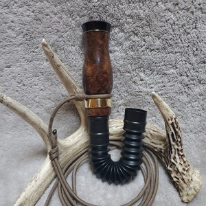May include: A wooden duck call with a black and gold finish, attached to a brown cord and a black flexible hose.