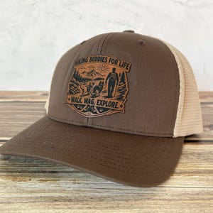 May include: A brown and tan trucker hat with a graphic that reads "Hiking Buddies for Life" and "Walk. Wag. Explore." The graphic features a person, two dogs, trees, and a sun. The hat has a curved brim and a mesh back.