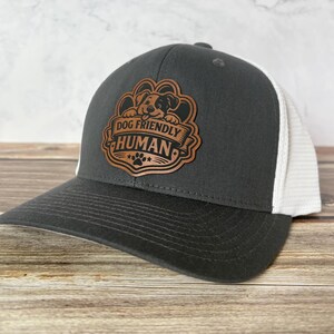 May include: A charcoal gray and white trucker hat with a curved brim. The hat features a brown leather patch with the words "DOG FRIENDLY HUMAN" and a dog illustration. The hat is on a wooden surface.