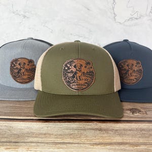 May include: Three baseball caps with a dog graphic. The caps are in gray, olive green, and navy blue. Each cap has a different city name: Clearwater, Dunedin, and Tampa, Florida. The caps have a mesh back and a curved brim.