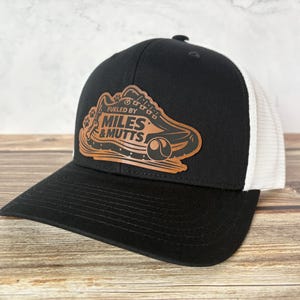 May include: Black baseball cap with a white mesh back and a curved brim. The cap features a brown leather patch with the words "Fueled by Miles & Mutts" and a graphic of a running shoe and paw prints.
