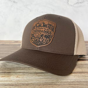 May include: A brown and tan trucker hat with a curved brim. The front panel features a leather patch with the words "Fueled by Coffee & Dogs" and illustrations of coffee cups and dogs.
