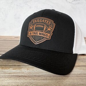 May include: Black and white trucker hat with a curved brim. The front panel features a brown leather patch with the words "TAILGATES & TAIL WAGS" and a football graphic. The hat has a mesh back.