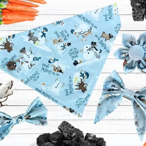 May include: A light blue pet bandana, bow tie, and flower accessory set. The items feature a winter-themed pattern with snowmen, dogs, and the text "Up to Snow Good". The bandana is fastened with a wooden button.