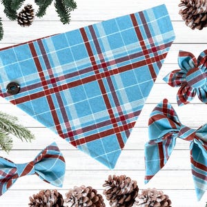May include: A collection of pet accessories in a blue and red plaid pattern. The set includes a bandana with a button closure, two bow ties, and a flower-shaped accessory. The accessories are arranged on a white wooden surface with pine cones and greenery.
