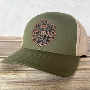 May include: Olive green and tan trucker hat with a leather patch. The patch features the text "PAWS PINTS TAILS & ALES" with a paw print, beer mug, and wheat design. The hat has a curved brim and a mesh back.
