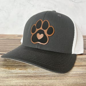 May include: A charcoal gray baseball cap with a white mesh back. The cap features a brown and black paw print design with a heart in the center. The brim is dark gray.