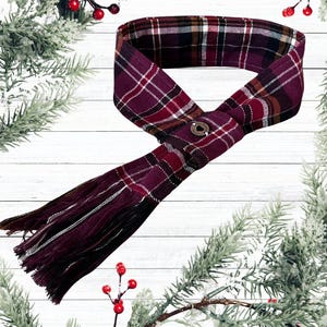 May include: A burgundy plaid scarf with fringe. The scarf features a pattern of burgundy, black, white, and gold. A metal clasp secures the scarf. The scarf is displayed on a white wooden background with holiday greenery.