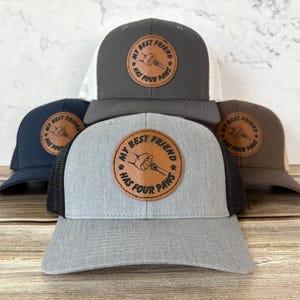 May include: A collection of trucker hats in various colors, including gray, navy, and brown. Each hat features a circular leather patch with the text "MY BEST FRIEND HAS FOUR PAWS" and a paw print design.