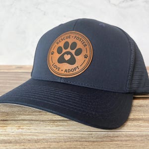May include: Navy blue baseball cap with a mesh back and a curved brim. The cap features a circular brown leather patch with the words "RESCUE FOSTER LOVE ADOPT" around a paw print and heart design.