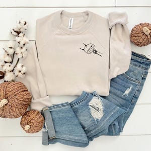 May include: A beige crewneck sweatshirt with a black graphic of a hand holding a paw, paired with distressed blue jeans. Decorative pumpkins and cotton stems complete the fall-themed ensemble.