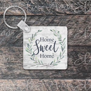 May include: A white square keychain with a woodgrain background and a green leafy wreath surrounding the text "Home Sweet Home".