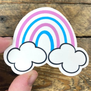 May include: A sticker featuring a rainbow with pink and blue stripes, and two white clouds with black outlines. The sticker is held against a wooden background.
