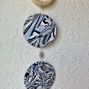 Puede incluir: Adorno de pared colgante con tres diseños circulares marmolados en negro, blanco y azul. Los círculos están unidos por una cuerda y cuentas rosas, con un anillo de madera en la parte superior. El patrón abstracto crea una estética única. Para decoración de pared.