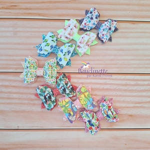 May include: A set of 7 colourful hair bows with various floral and animal prints. The bows are made of fabric and have a layered design. The bows are arranged in a row on a wooden surface. The text "Bouclette par Karal Prevost" is visible in the image.