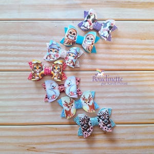 May include: A set of 10 colourful hair bows featuring various animal designs, including owls, deer, tigers, and bunnies. The bows are made of fabric and have a glitter finish. The bows are arranged in a row on a wooden surface. The text "Bouclinette par Karine Prévost" is visible in the image.