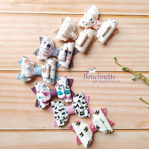 May include: A set of eight colourful hair bows with a cat print design. The bows are made of fabric and have a glittery finish. The bows are arranged in a semi-circle on a wooden surface.