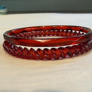 May include: Two stacked bangles made of translucent deep red material. The top bangle features a smooth, faceted outer edge, while the bottom bangle has a distinct, continuous spiral or twisted rope-like texture throughout its circumference.