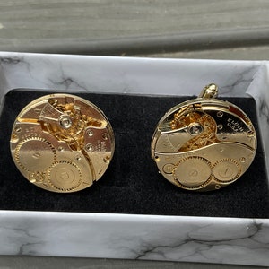 May include: A pair of gold-toned circular cufflinks crafted from vintage Elgin U.S.A. watch movements, featuring exposed gears, springs, and mechanical components. The cufflinks are displayed inside a white box with a grey marble pattern.