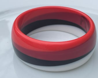 May include: A thick, circular bangle bracelet featuring a smooth, glossy finish. The design consists of three distinct, horizontal color bands in vibrant red, solid black, and crisp white, arranged in a stacked pattern around the entire circumference.