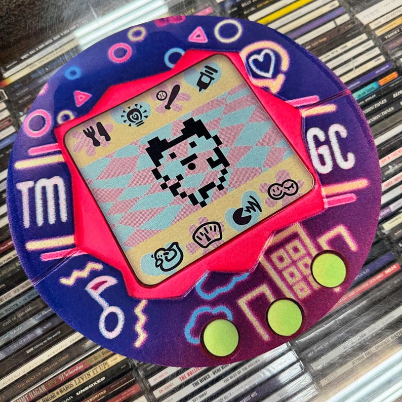 K&ouml;nnte beinhalten: Ein Retro-Handheld-Digital-Pet-Spiel mit einem lebendigen Design. Das Ger&auml;t hat einen pinkfarbenen Rahmen, einen Bildschirm, der einen pixeligen Charakter anzeigt, und neongr&uuml;ne Kn&ouml;pfe. Das &auml;u&szlig;ere Geh&auml;use ist blau mit rosa und wei&szlig;en Akzenten, und die Buchstaben "TM" und "GC" sind sichtbar.