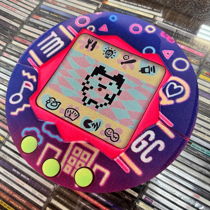 K&ouml;nnte beinhalten: Ein rundes, lila und pinkfarbenes Retro-Handheld-Spielger&auml;t mit einem pixeligen Charakter auf dem Bildschirm. Das Ger&auml;t hat neongr&uuml;ne Kn&ouml;pfe und verschiedene Symbole. Der Hintergrund ist eine Sammlung von CDs.