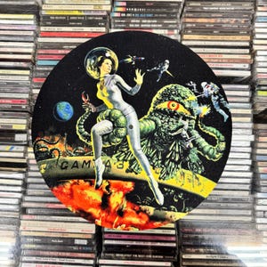 May include: A circular graphic featuring a retro sci-fi scene with a woman in a spacesuit, a large green monster, and spaceships. The image has a black background with the text "GAMMA 3" and is surrounded by stacks of CDs.
