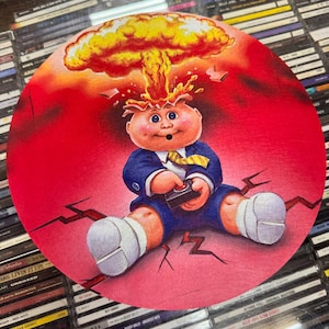 May include: A round, red coaster with a cartoon image of a baby with a mushroom cloud exploding from its head. The baby is wearing a blue suit and a yellow tie. The baby is sitting on a cracked surface with lightning bolts.