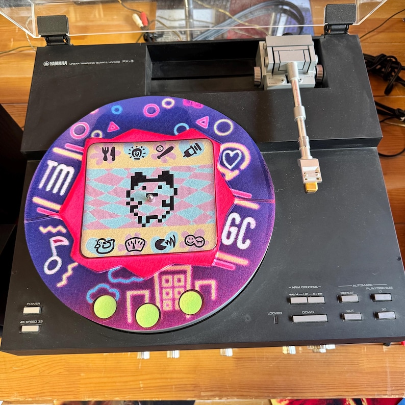 K&ouml;nnte beinhalten: Schwarzer Yamaha PX-3 Plattenspieler mit einer individuellen Schallplatte mit einem pixeligen Charakterdesign. Die Schallplatte hat ein rosa-violettes Farbschema mit einer Stadt und geometrischen Formen.
