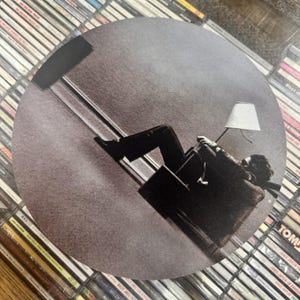 May include: Circular album art featuring a grayscale image of a person reclining in a chair with a lamp. The person is positioned at an angle, creating a dynamic composition. The background is a collection of CDs.