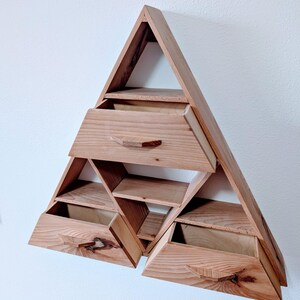 May include: A wooden, triangular wall shelf with multiple compartments and drawers. The shelf is made of light-colored wood and features a geometric design. The drawers have small, wooden handles. The shelf is mounted on a white wall.