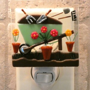 May include: A green glass night light shaped like a wheelbarrow with brown, orange, and yellow flowers and green leaves. The wheelbarrow is filled with brown dirt and has a silver wheel. The night light is plugged into an outlet.