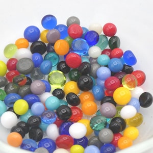 May include: A collection of small, round glass beads in a variety of colors including red, blue, yellow, green, orange, black, white, and gray. The beads have a smooth, polished finish and are scattered together in a clear glass bowl.