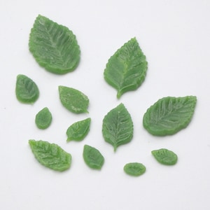 May include: A collection of small, green, leaf-shaped embellishments. The leaves vary in size and shape, with detailed veining visible on each. They are arranged on a white surface, suggesting a craft or decorative theme.