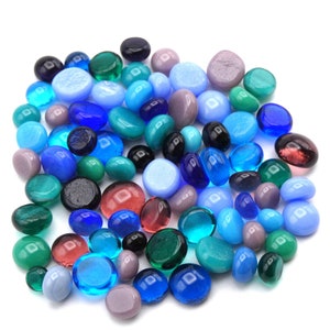 May include: A collection of colorful glass pebbles in shades of blue, green, and purple. The smooth, rounded stones vary in size and opacity, creating a visually appealing assortment. Some pebbles are transparent, while others are opaque.