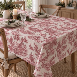 Puede incluir: Un mantel rectangular con un patrón toile de jouy en rojo y blanco que muestra escenas pastorales detalladas. La tela cubre una mesa de madera, la cual está dispuesta con platos blancos, servilletas dobladas, cristalería y una jarra.