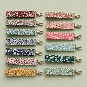 Puede incluir: Una colección de catorce llaveros de tela tipo pulsera dispuestos en dos columnas. Cada llavero presenta un patrón floral único en varios colores, incluyendo rojo, azul, verde, rosa y amarillo, y está unido a un mosquetón de metal plateado.