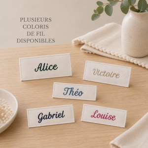 Può includere: Cinque etichette rettangolari in tessuto color crema con nomi ricamati in vari colori: Alice in verde scuro, Victoire in beige, Théo in blu, Gabriel in blu scuro e Louise in rosa. Testo: PLUSIEURS COLORIS DE FIL DISPONIBLES.