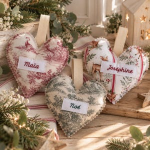 Könnte beinhalten: Drei herzförmige Stoffanhänger mit beigen Schlaufen. Einer zeigt ein rotes Toile-Muster mit dem Namen Maïa, einer ein grünes Toile-Muster mit dem Namen Noé und einer einen Wald-Hirsch-Druck mit dem Namen Joséphine auf einem weißen Etikett.