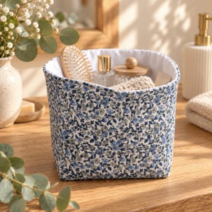 Op de afbeelding: Een stoffen opbergmand met een blauw-wit bloemenpatroon en een witte binnenvoering. De mand is gevuld met diverse badkameraccessoires, waaronder een haarborstel, een glazen flesje en een klein bakje met een houten deksel.