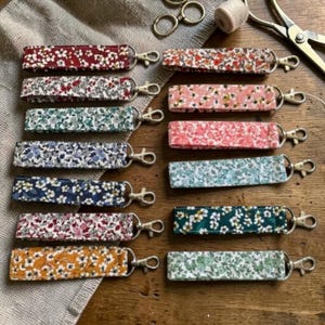 Puede incluir: Una colección de llaveros de tela con estampados florales en varios colores, como rojo, rosa, azul y amarillo. Cada llavero rectangular tiene un cierre metálico. Los llaveros están dispuestos sobre una superficie de madera.