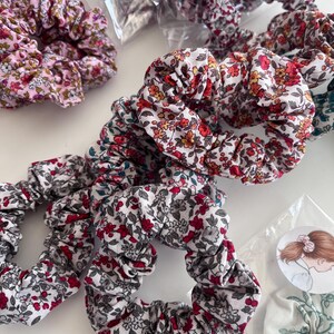 K&ouml;nnte beinhalten: Eine Sammlung von Stoff-Haargummis mit verschiedenen Blumenmustern in Rot-, Rosa-, Orange- und Gr&uuml;nt&ouml;nen auf wei&szlig;em Hintergrund. Einige Haargummis liegen lose, andere sind in durchsichtigen Plastikt&uuml;ten mit K'Alice Deco-Etiketten verpackt.