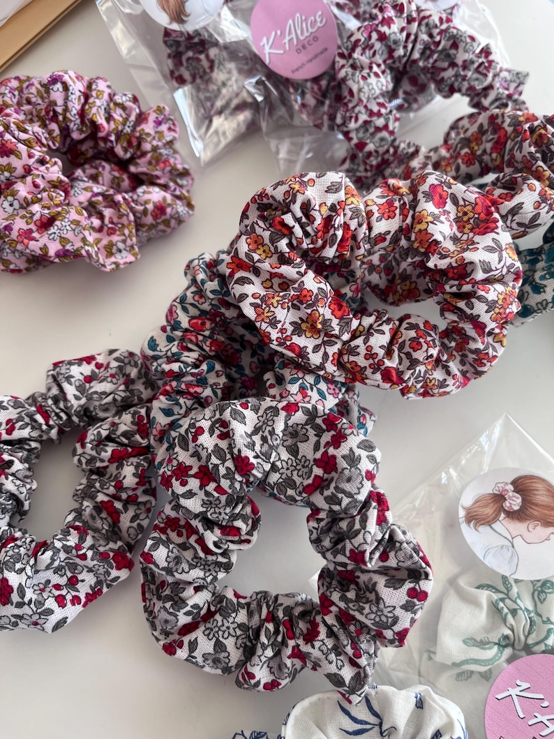 K&ouml;nnte beinhalten: Eine Sammlung von Stoff-Haargummis mit verschiedenen Blumenmustern in Rot-, Rosa-, Orange- und Gr&uuml;nt&ouml;nen auf wei&szlig;em Hintergrund. Einige Haargummis liegen lose, andere sind in durchsichtigen Plastikt&uuml;ten mit K'Alice Deco-Etiketten verpackt.