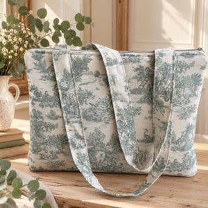 May include: A rectangular tote bag made of off-white fabric featuring a repeating sage green toile de jouy pattern depicting pastoral scenes with trees, figures, and birds. The bag includes two long shoulder straps and a top zipper closure.